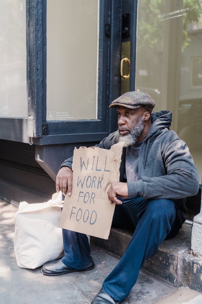 An adult man sitting on sidewalk holding a cardboard sign, addressing social issues.